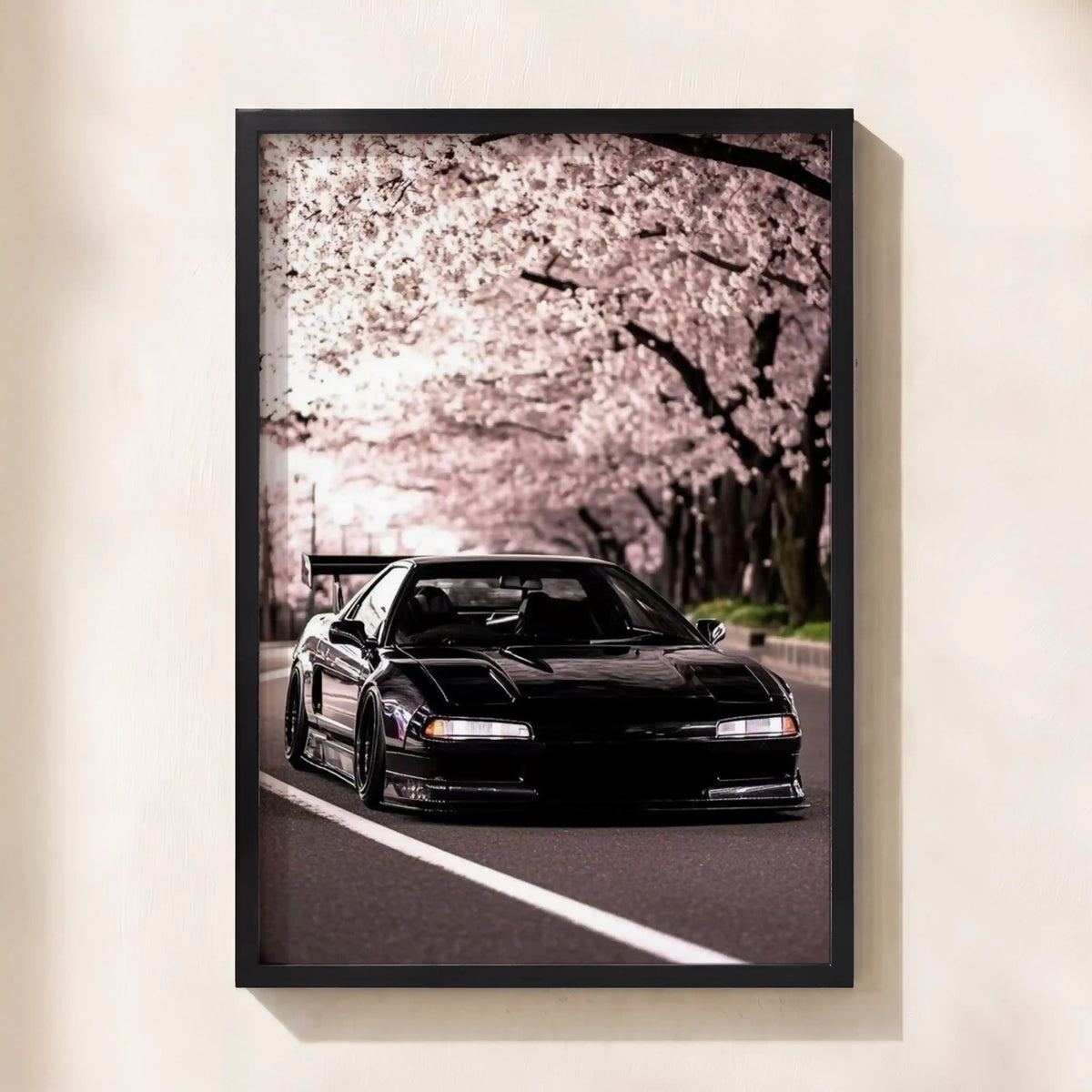 A framed vertical art print featuring a black sports car driving on a road beneath blooming cherry blossom trees, displayed on a light beige wall.