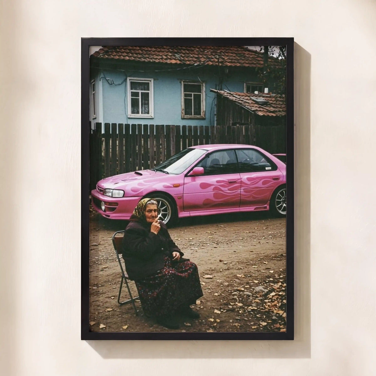 A framed photographic print hanging on a light beige wall. The print shows a woman sitting on a chair in front of a pink car and a blue house.