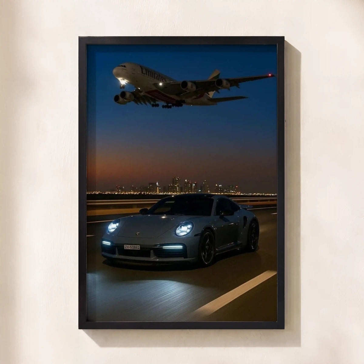 A framed vertical art print depicting a gray sports car on a highway at dusk with an Emirates airplane flying overhead, mounted on a light beige wall.