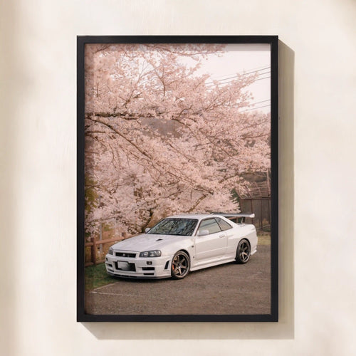 A framed vertical art print featuring a white sports car parked under pink cherry blossoms, mounted on a light beige wall.