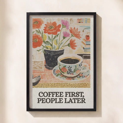 A framed art print featuring a vintage-style illustration of flowers in a vase and a cup of coffee, with the text 'COFFEE FIRST, PEOPLE LATER' at the bottom, displayed on a light beige wall.