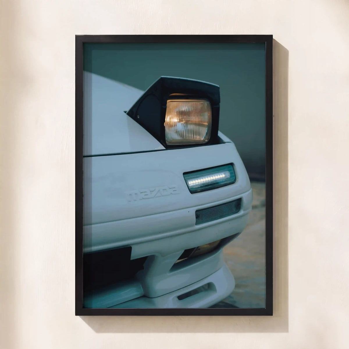 A framed photographic print of a white Mazda RX-7 car front end, mounted on a light beige wall.