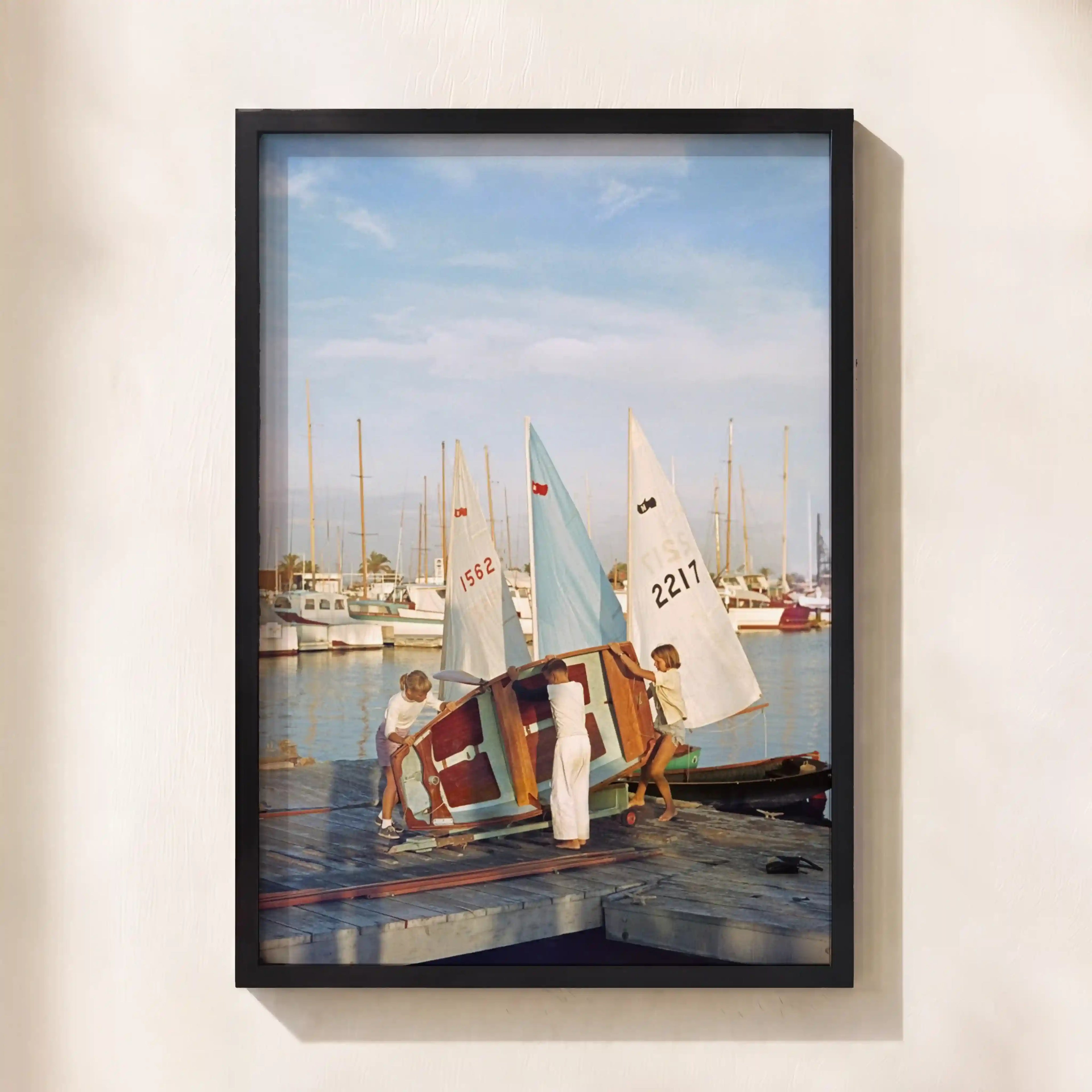 A framed photographic print of a marina scene, showing children handling a small sailboat on a dock with other boats in the background, mounted on a light beige wall.