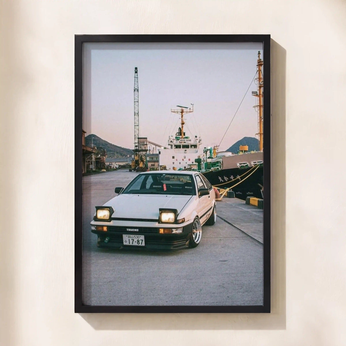 A framed photographic print of a white Toyota Corvette parked at a harbor, mounted on a light beige wall.
