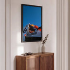 A framed vertical art print featuring a motorcycle racer in a white Marlboro helmet against a bright blue sky, displayed on a white wall above a wooden sideboard with a vase of eucalyptus.