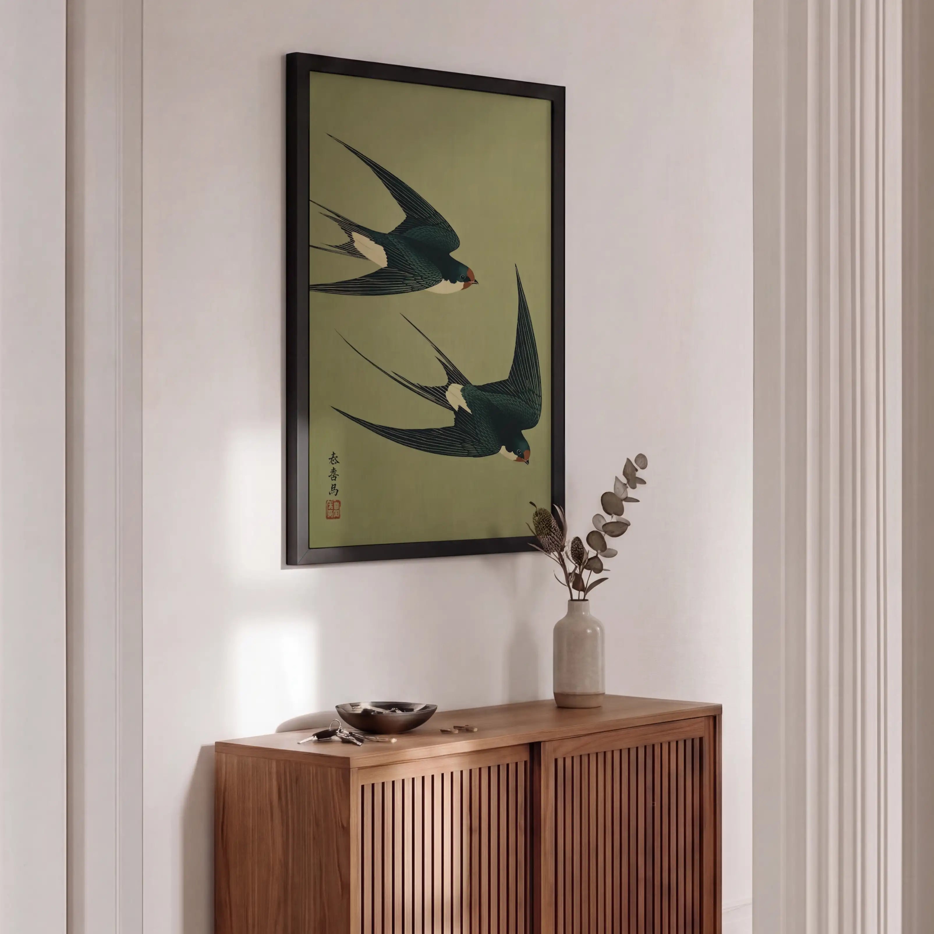A framed art print depicting two flying swallows on an olive green background, hanging on a white wall above a wooden credenza in a modern, sunlit room.