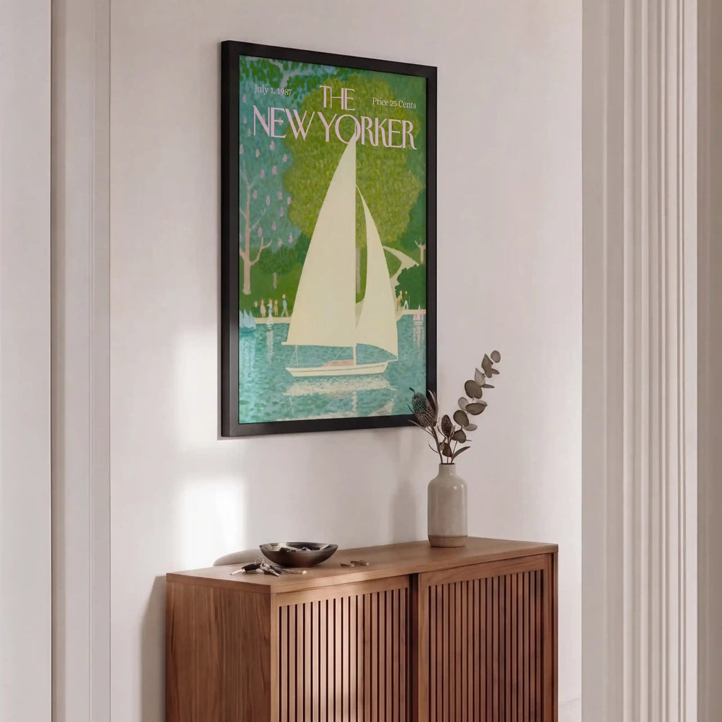 A framed New Yorker magazine cover featuring a sailboat illustration in green, teal, cream, and blue tones, displayed on a white wall above a wooden credenza in a styled interior.