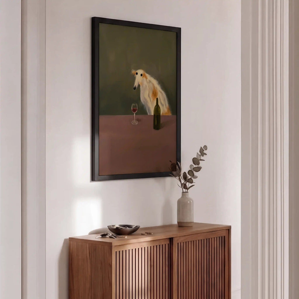 A framed art print featuring a white dog at a table, hanging on a white wall above a wooden credenza in a modern interior setting.
