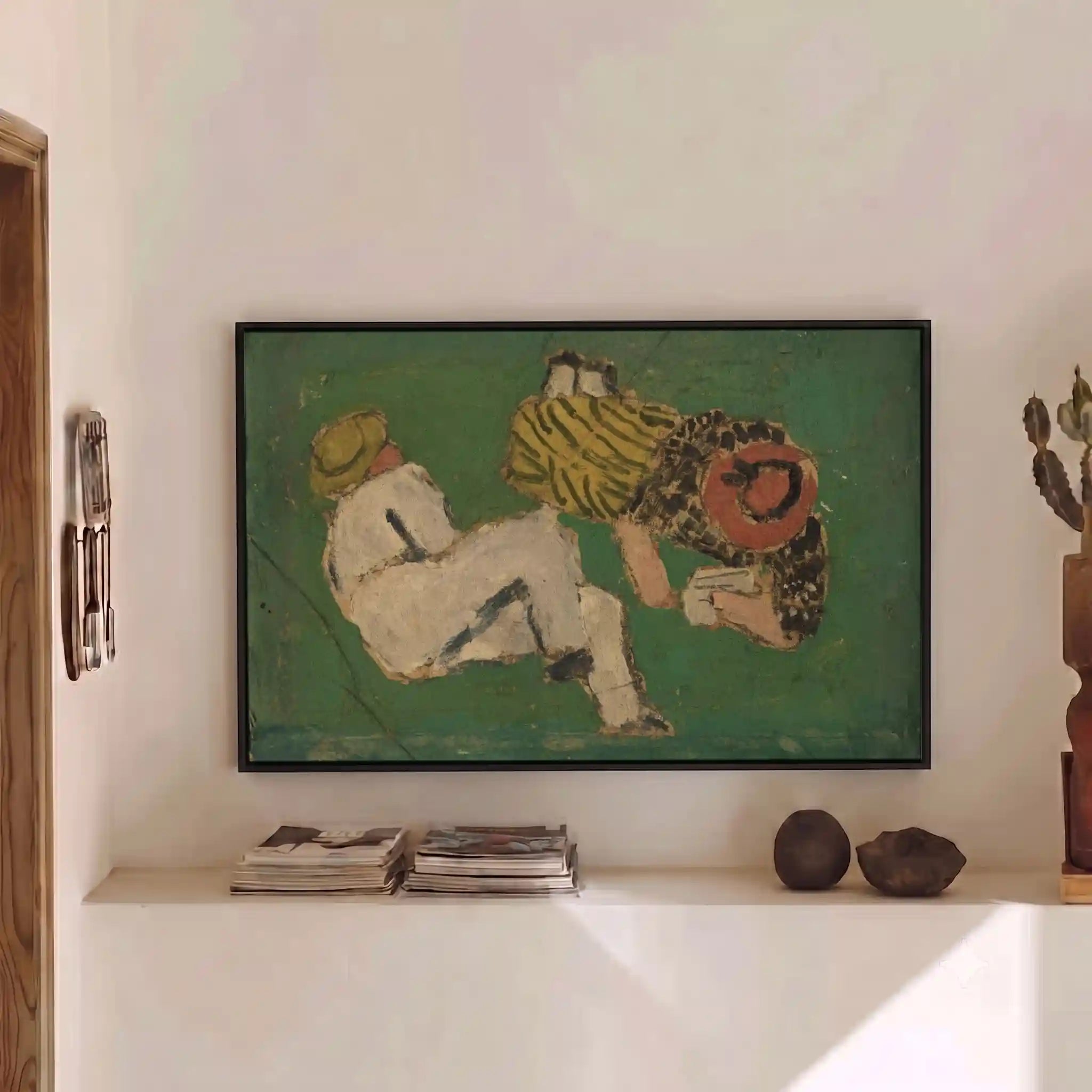 A framed art print depicting two figures in a green-toned painting, displayed on a white wall above a shelf with books and decorative objects.