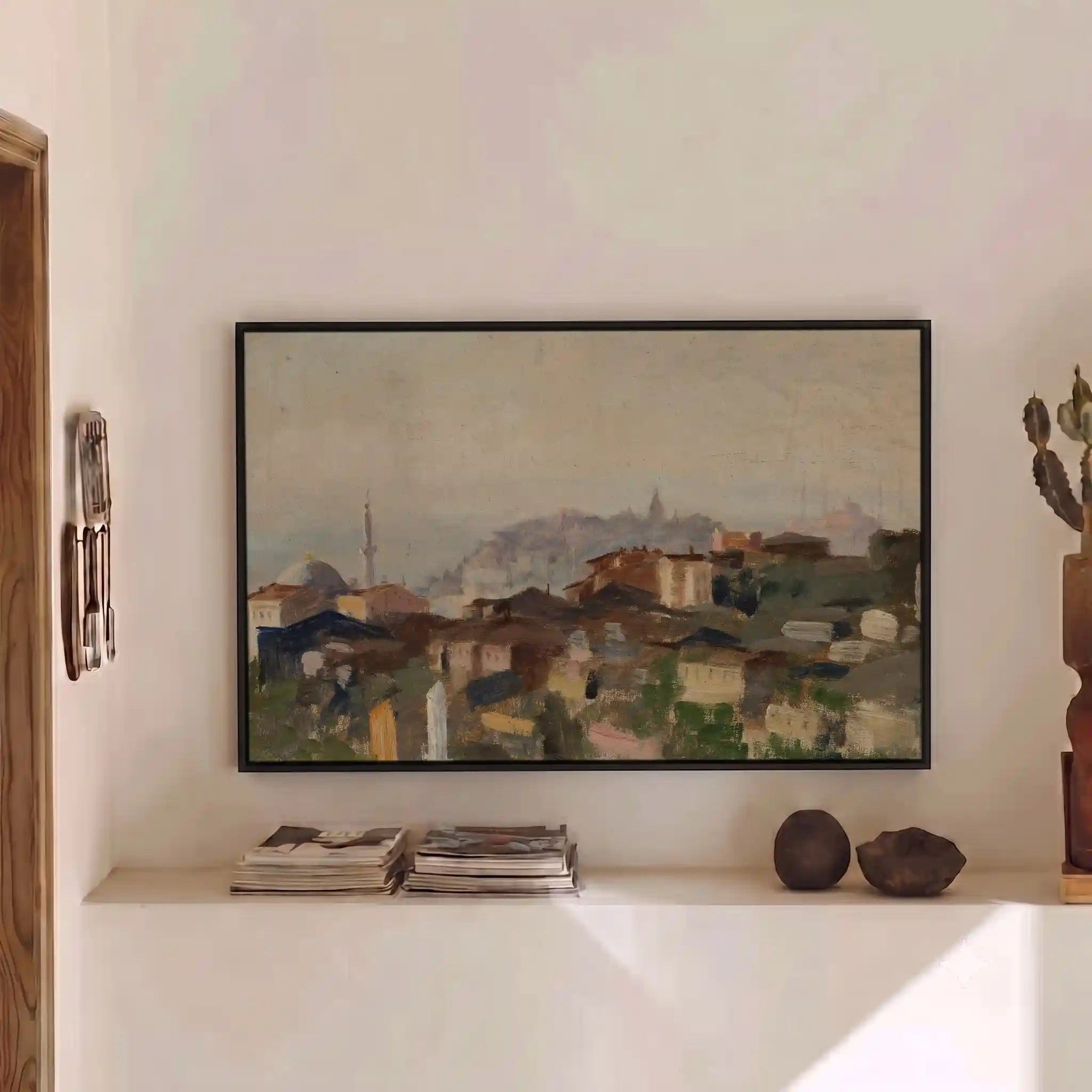 A framed landscape painting of a hillside village with muted earth tones, displayed on a white wall above a shelf with books and decorative objects.