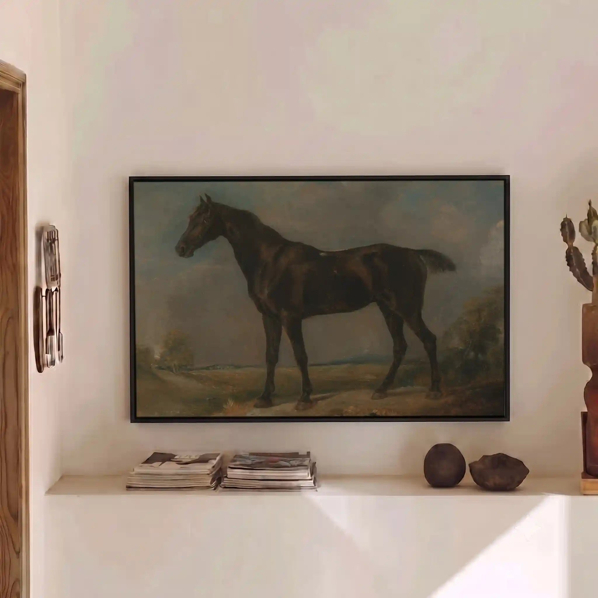 A framed classical painting of a dark horse standing in a landscape, displayed on a white wall above a shelf with books and decorative objects.