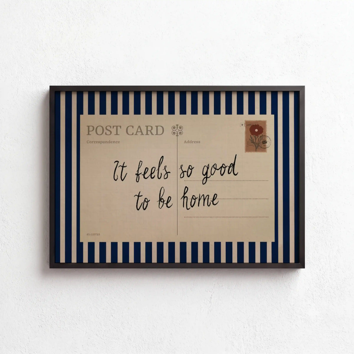 A framed art print featuring a vintage postcard design with navy blue and cream vertical stripes, a cream postcard center with the handwritten text 'It feels so good to be home', and a black frame, mounted on a textured white wall.