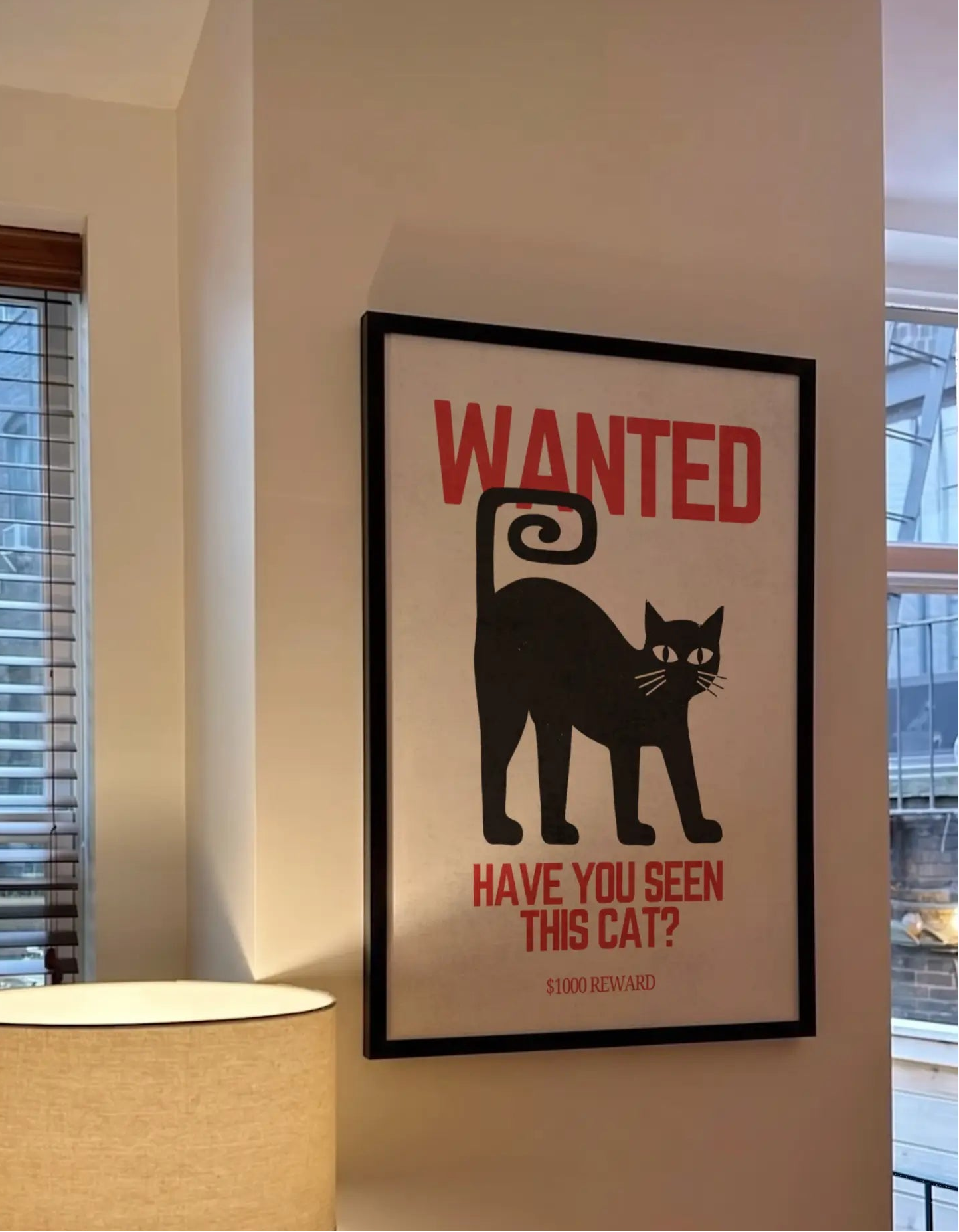 Framed 'Wanted' poster with a black cat on a wall in a room with a lamp and window blinds.