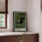 Framed artwork of a dachshund on a wall next to a window.